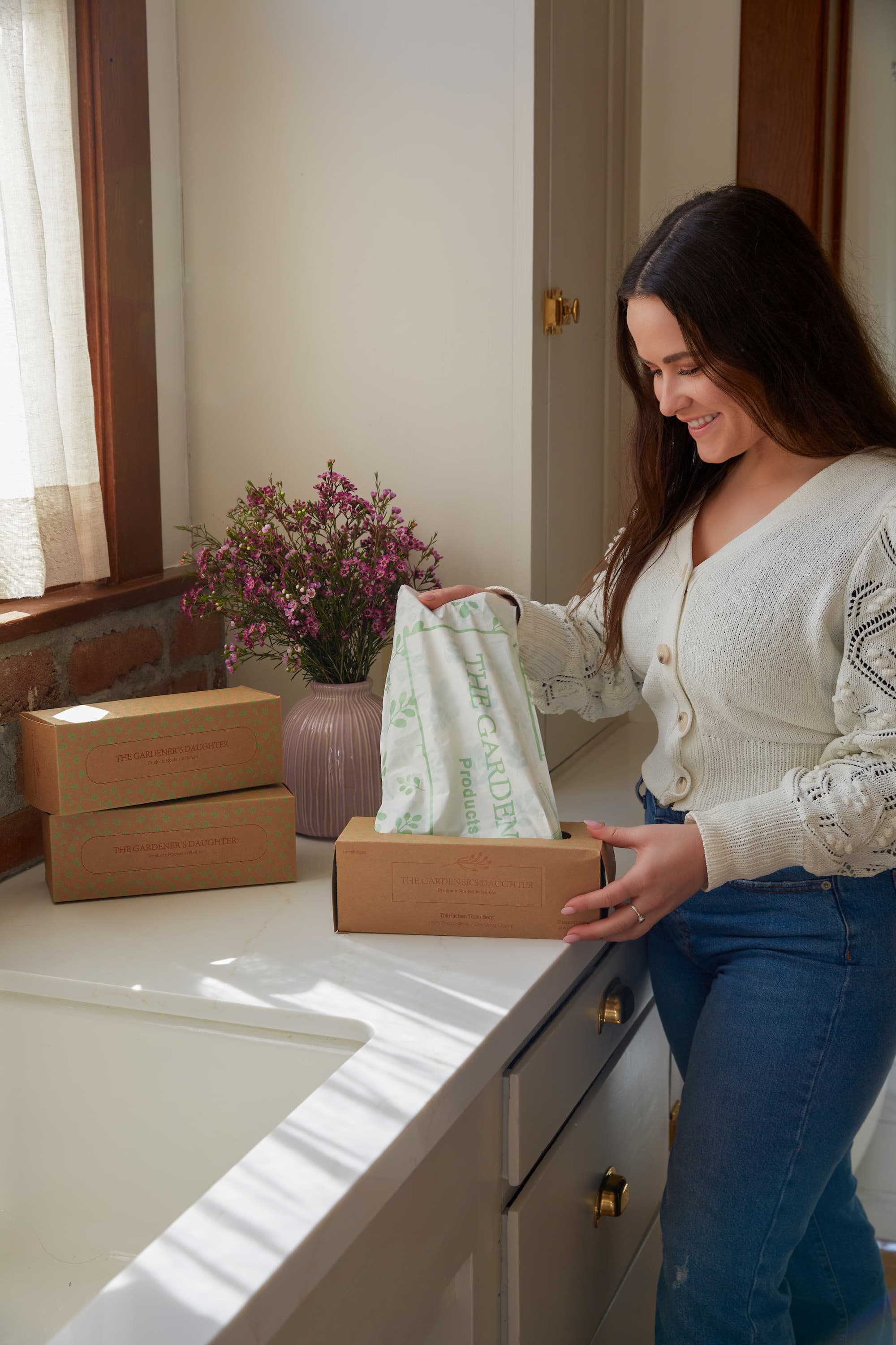 Woman smiling in a kitchen holding a The Gardener's Daughter compostable trash bag snext to pink flowers.